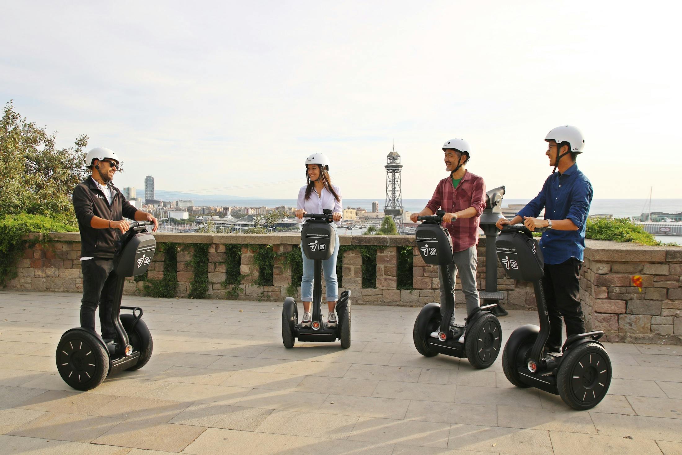 四个戴头盔的人在一个有城市景观和塔楼背景的铺面区域乘坐赛格威。