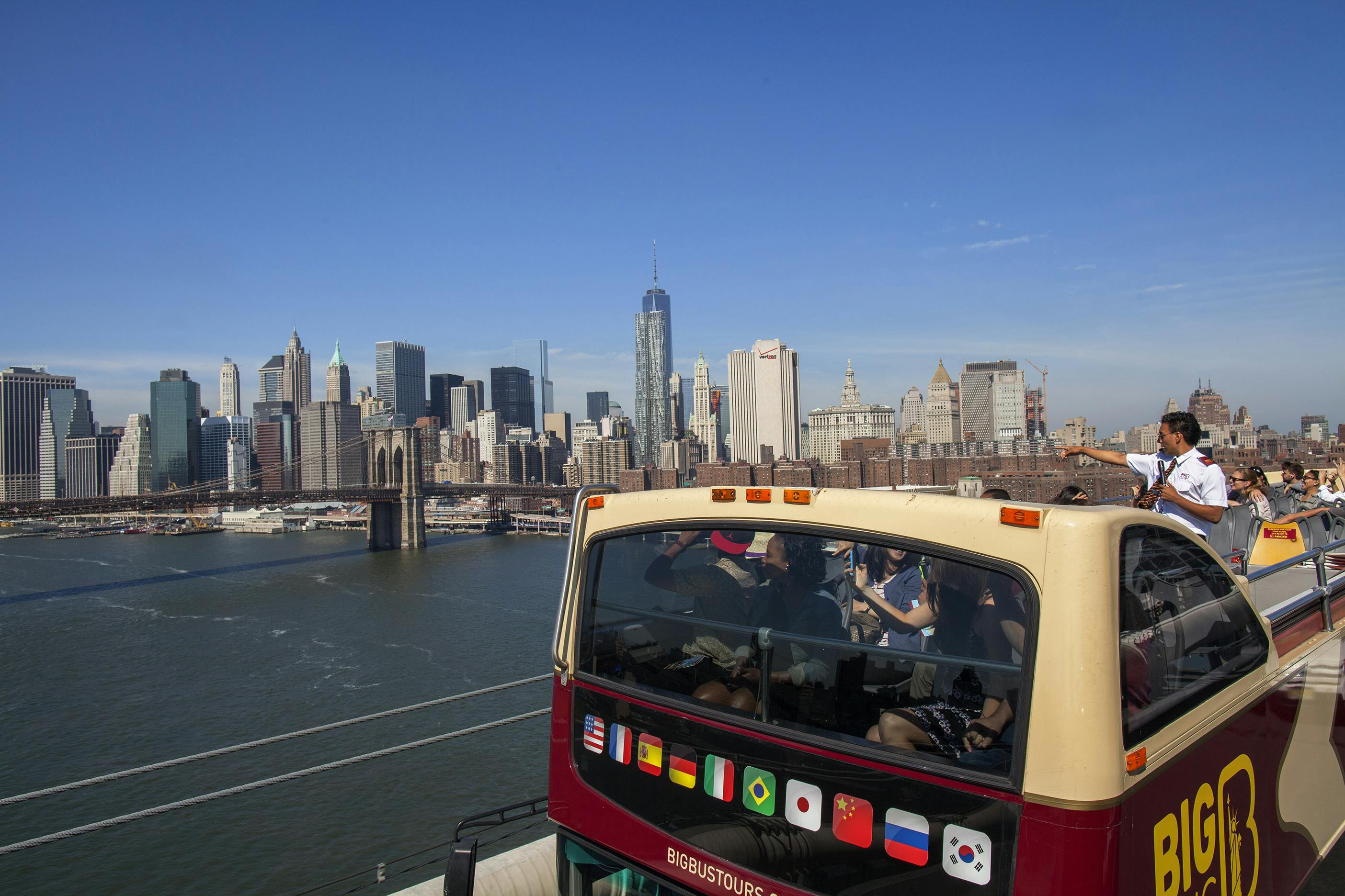 A double-decker tour bus crosses a bridge with passengers viewing the New York City skyline and Brooklyn Bridge in the background.