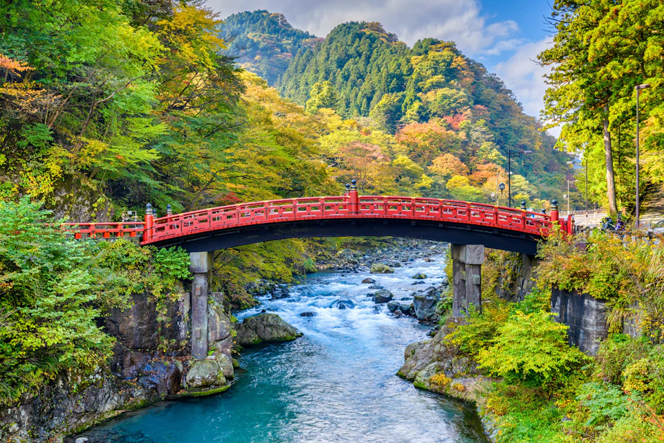 A traditional red bridge spans a clear river, surrounded by lush green and autumnal trees under a partly cloudy sky.