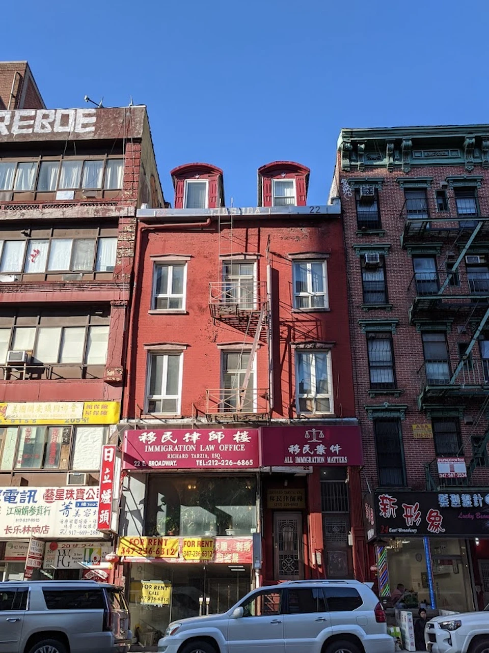 A red brick building with a fire escape, surrounded by other buildings. Signs in different languages are visible at street level.