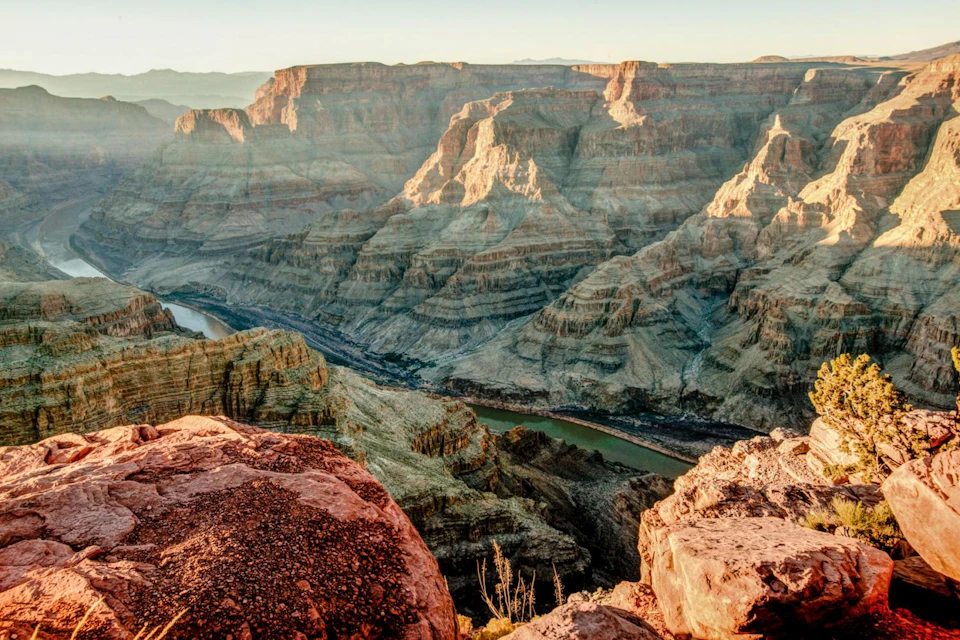 Rocky canyon landscape with layered, sunlit cliffs and a winding river at the bottom. Sparse vegetation in the foreground.