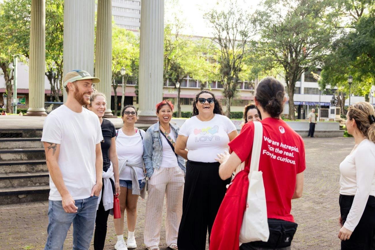 A group of people standing outdoors near columns, smiling and talking, with greenery and buildings in the background.