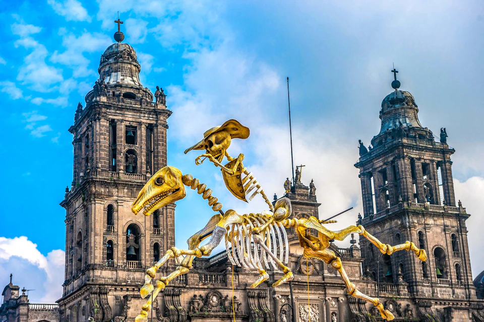 Golden skeletal dinosaur sculpture in front of historic stone buildings with towers, against a partly cloudy blue sky.