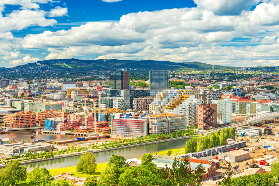 Colorful cityscape with modern buildings, a river, and surrounding greenery under a partly cloudy sky with distant hills.