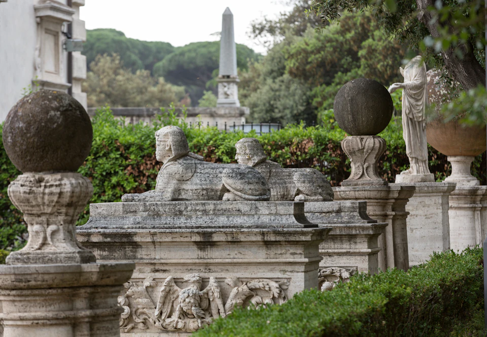 Outdoor scene featuring stone sphinx statues with human heads, an obelisk in the background, and lush greenery surrounding them.