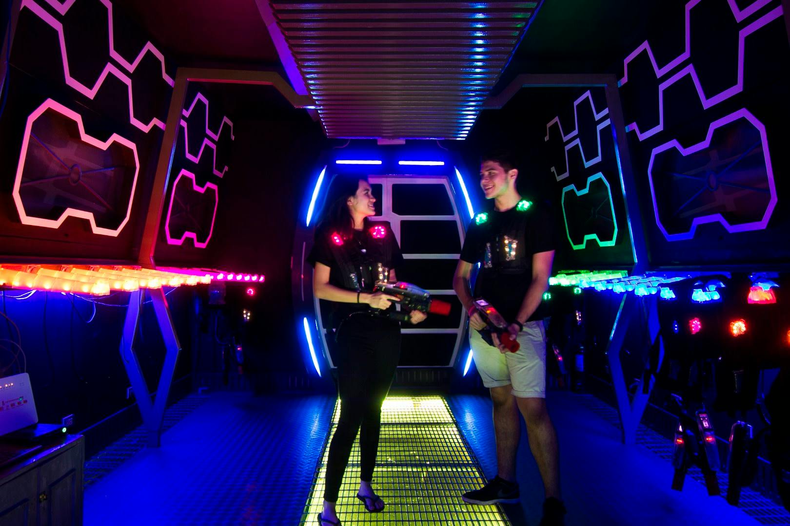 Two people wearing laser tag vests and holding laser guns stand in a neon-lit arena, smiling at each other.