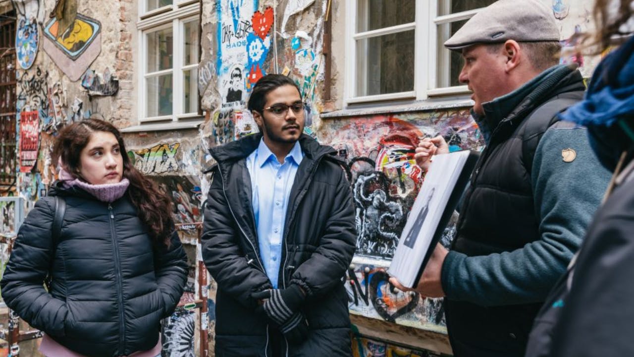 Three people stand in front of a graffiti-covered wall; one person holding a photo.