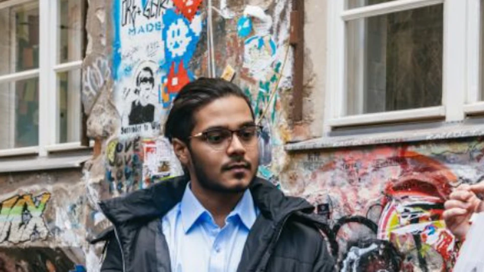 Three people stand in front of a graffiti-covered wall; one person holding a photo.