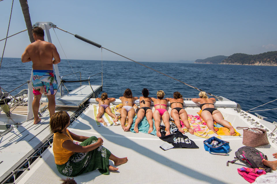People relaxing and sunbathing on a boat deck at sea, with the coastline and hills visible in the distance.