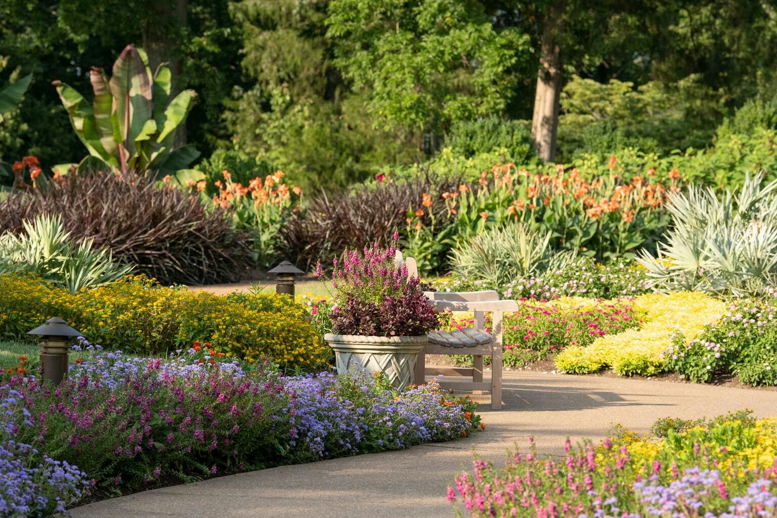 Zahrada s barevnými květinami, dřevěná lavička s polštáři a velký květináč na dlážděné cestě pod zelenými stromy.