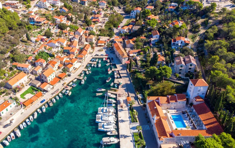 Aerial view of a coastal town with boats docked in turquoise water, red-tiled roof buildings, and lush greenery.