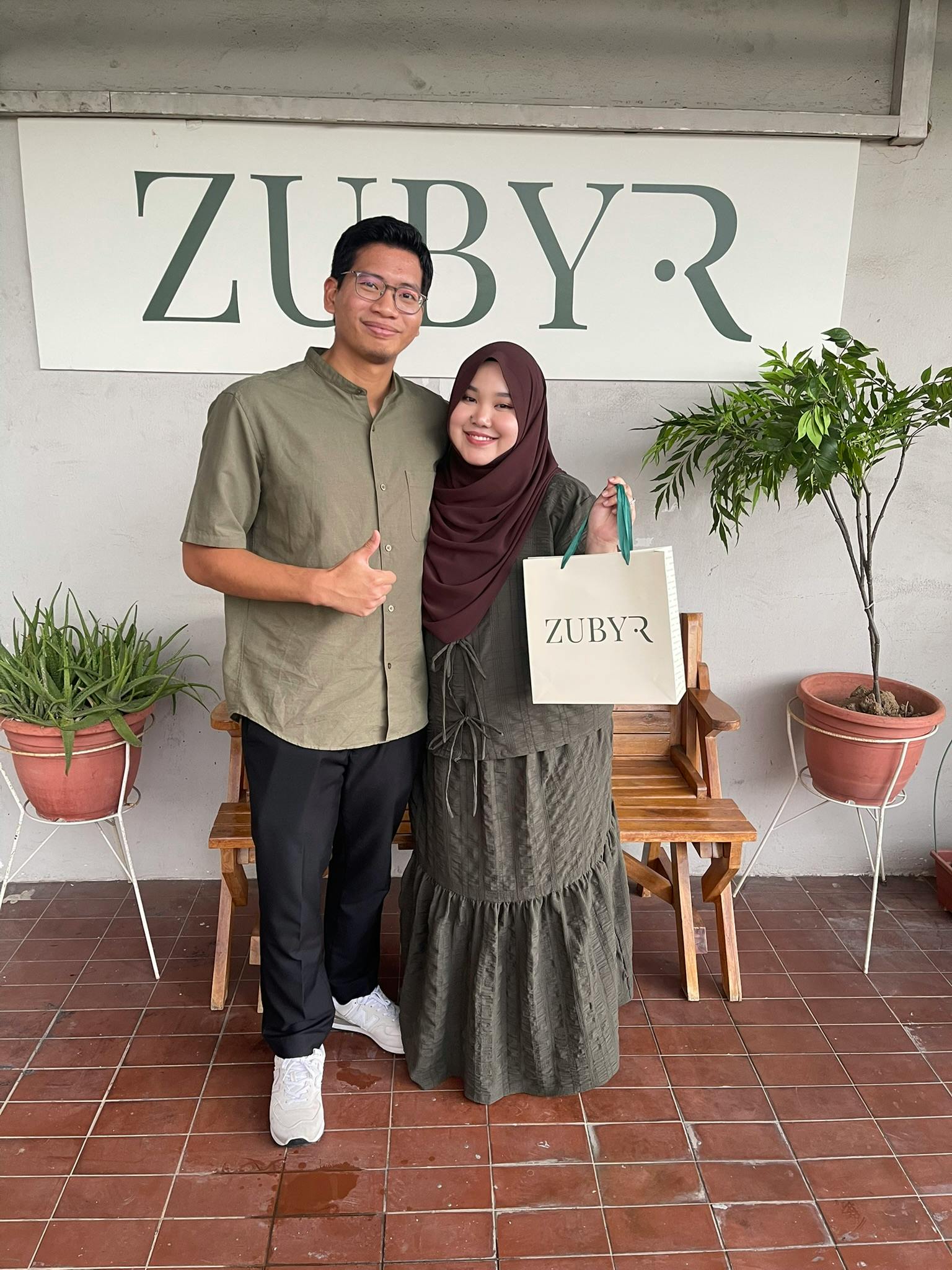 Two individuals stand smiling in front of a sign reading 'ZUBYR,' with one holding a branded shopping bag. Plants surround them.