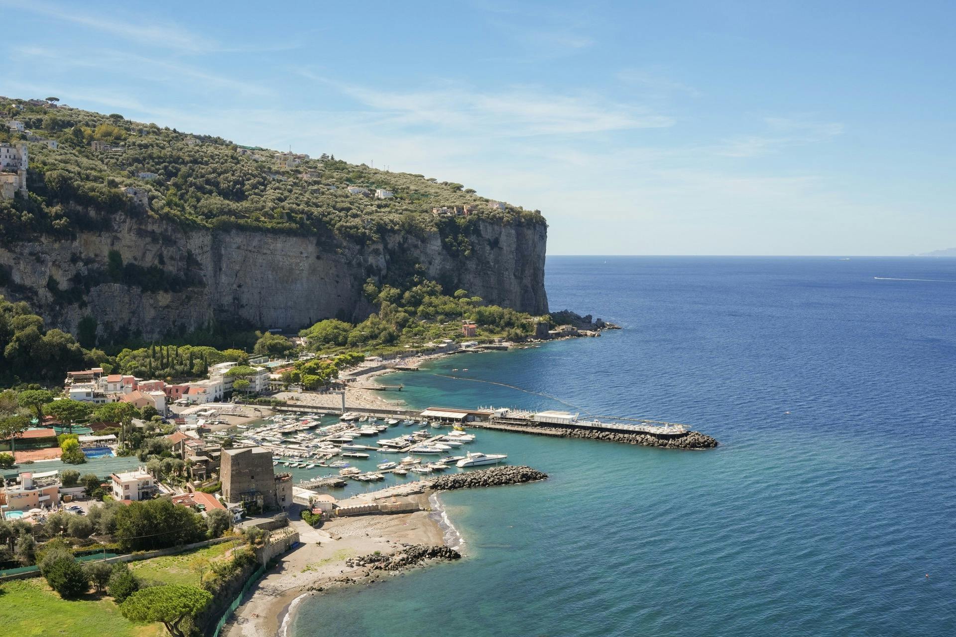 澄んだ空の下で青い海を見下ろすマリーナ、ボート、建物、崖のそばがある海岸沿いの町。