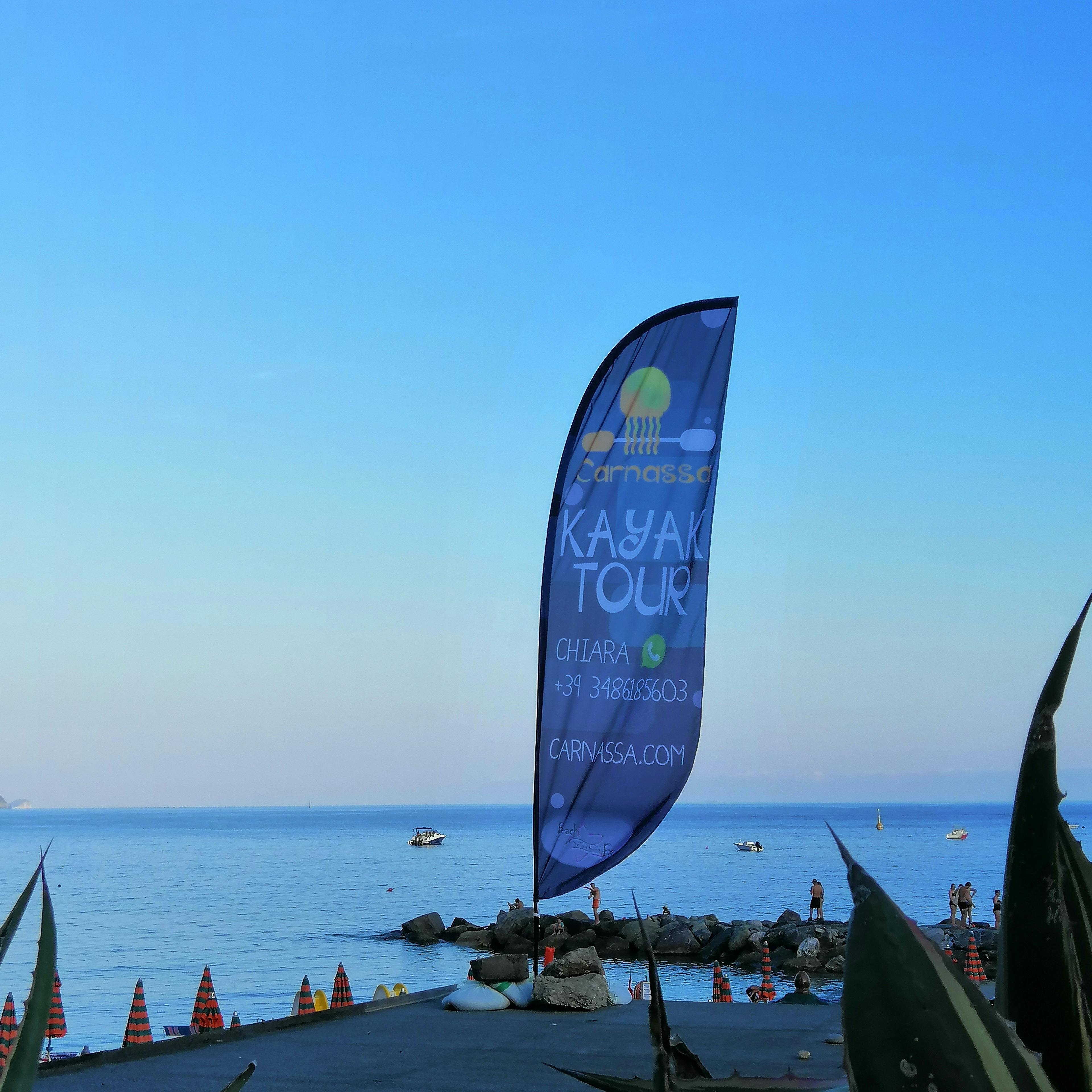 Una bandera que anuncia excursions en caiac es troba en una riba rocosa prop d'un mar tranquil, amb els vaixells i la gent de fons.