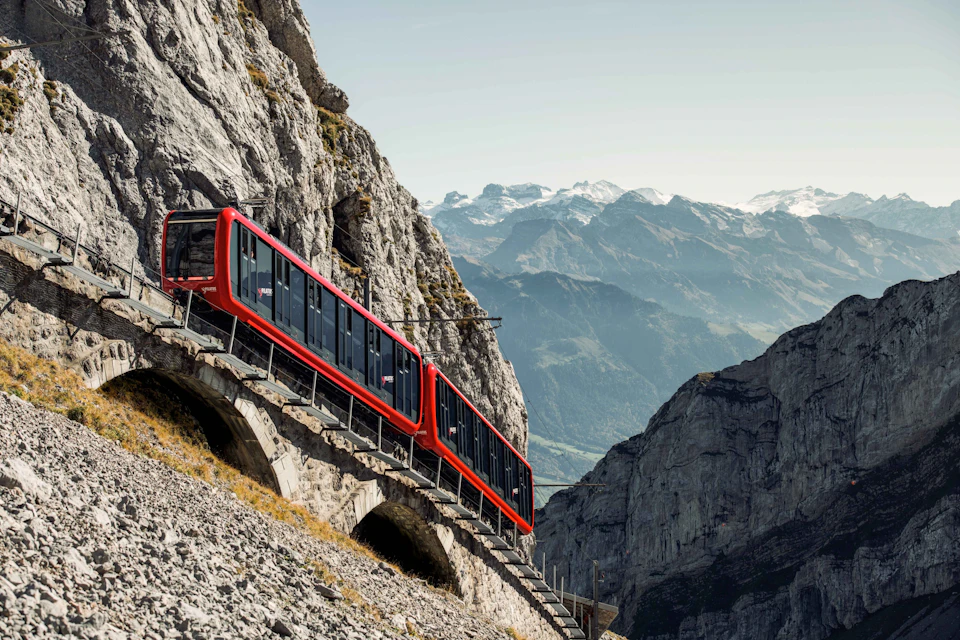 A red train traveling on a mountainside railway with rugged peaks in the background under a clear sky.