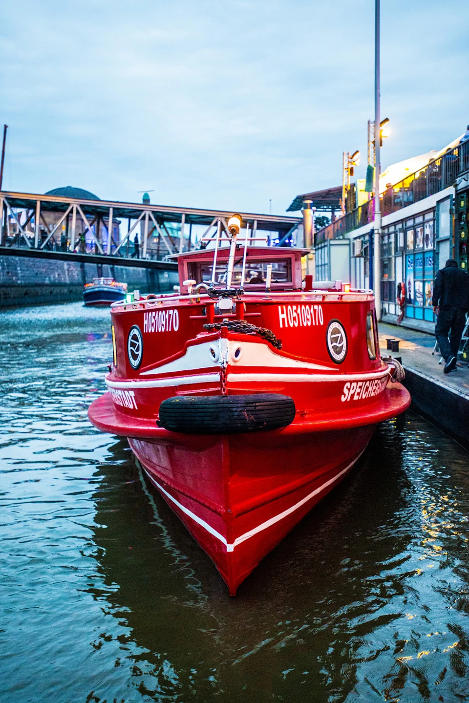 A red boat docked in a canal with a bridge, buildings, and people in the background.