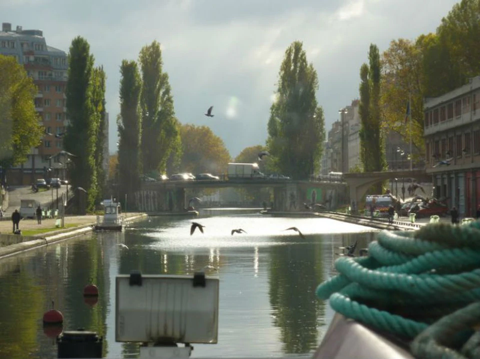A canal lined with trees and buildings, featuring a bridge, birds in flight, and a boat rope in the foreground.