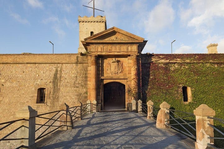 Stone fortress entrance with an arched doorway, surrounded by a vine-covered wall, and a bridge leading towards it.