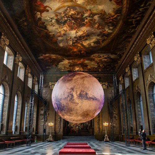 Påkostad hall med välvda fönster, ett detaljerat målat tak, en stor upphängd planetmodell och en person som tar bilder.