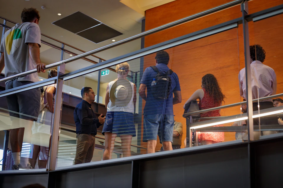 A group of people is gathered indoors near a railing, with one person speaking to the others, in a modern building setting.