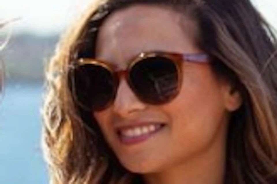 Three women in sweaters and jeans, wearing sunglasses, smile and hold drinks on a boat with water and a sailboat in the background.