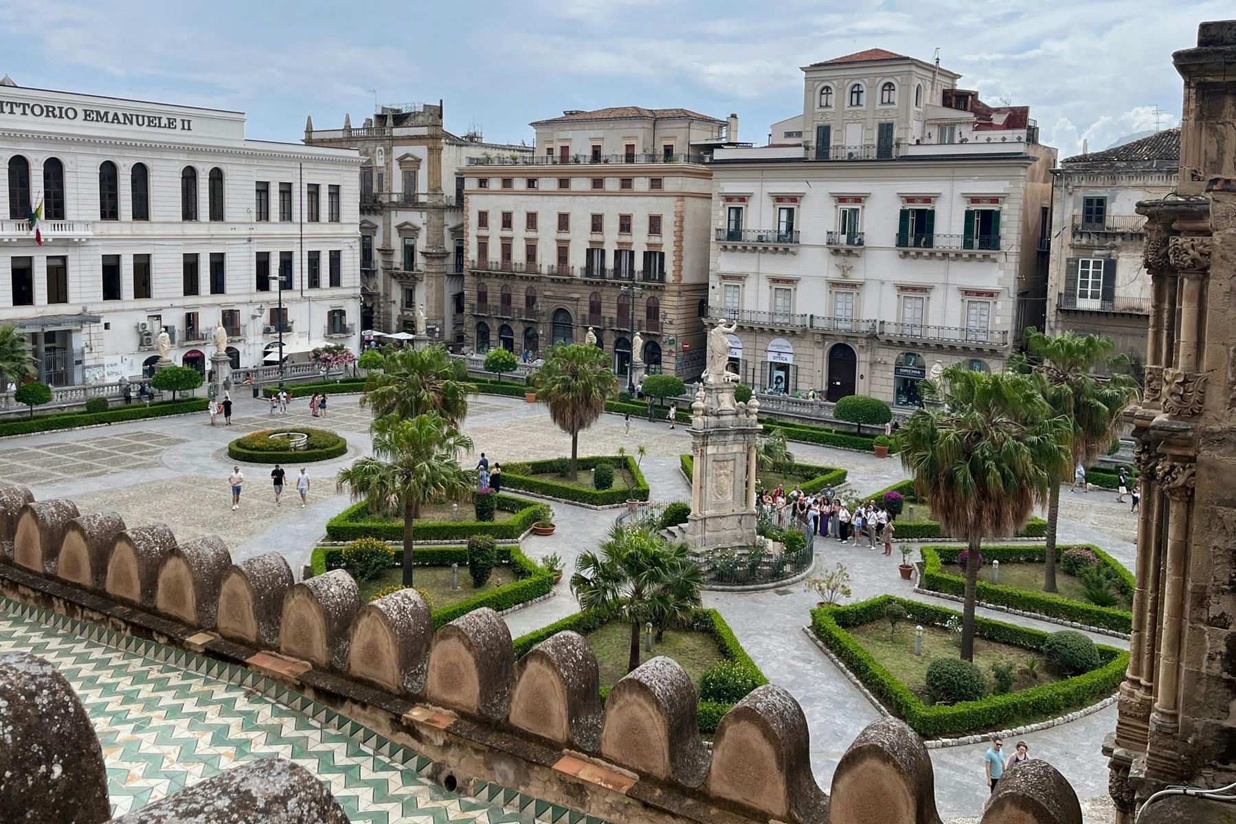 건물과 야자수로 둘러싸인 중앙 동상이 있는 광장을 걷는 사람들.