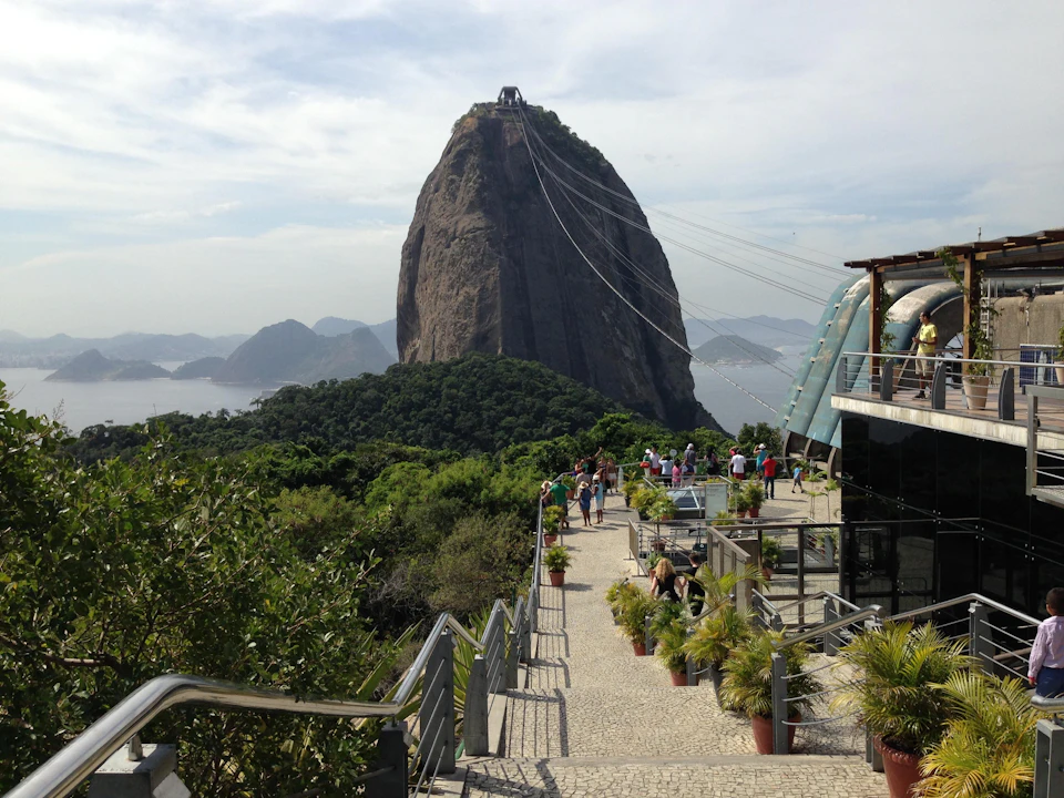 A large rock formation with a viewing platform and people walking along a pathway surrounded by greenery and railings.