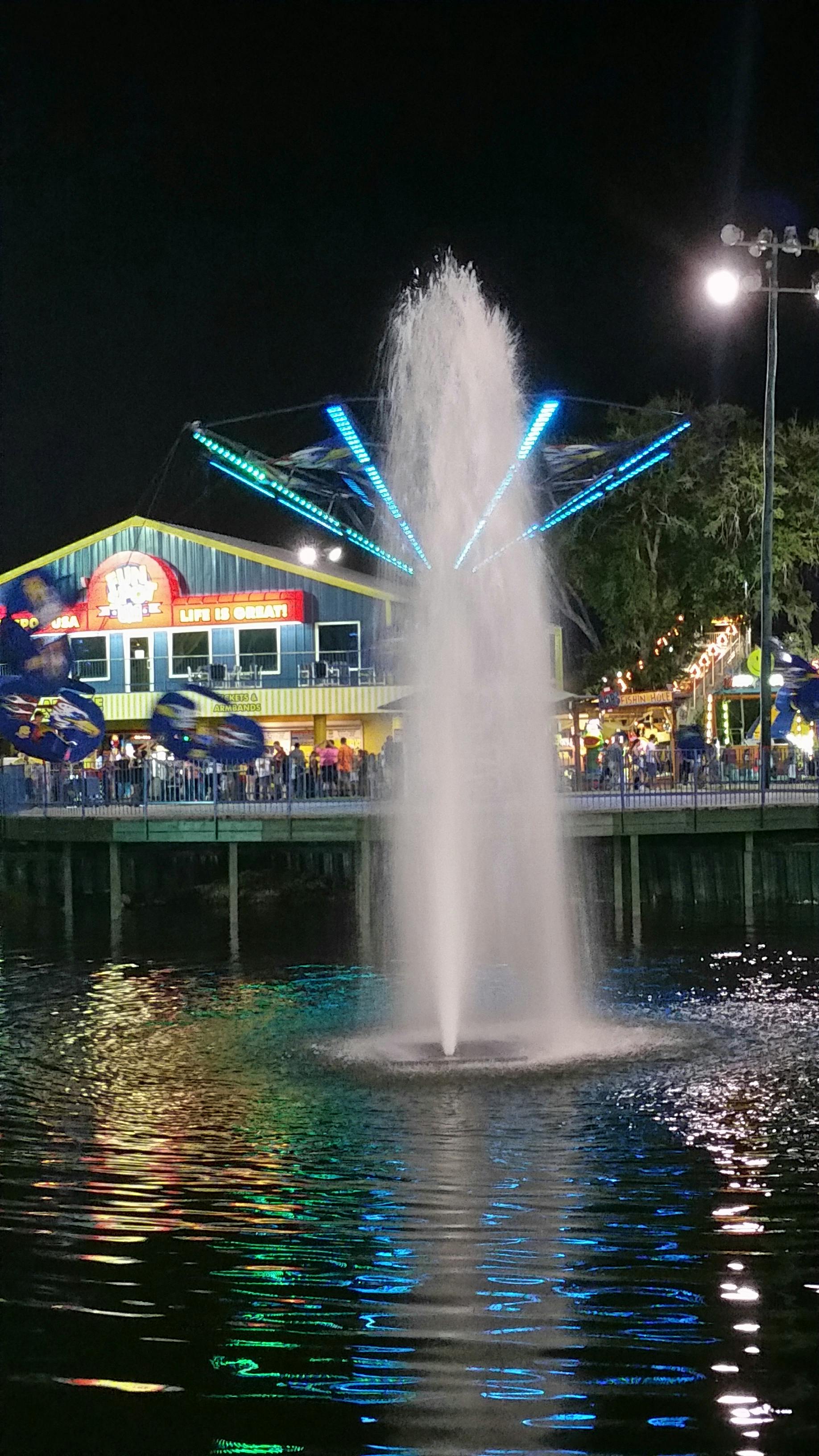 夜间游乐园，前景有一个点亮的喷泉，色彩缤纷的旋转游乐设施，建筑背景有明亮的灯光。