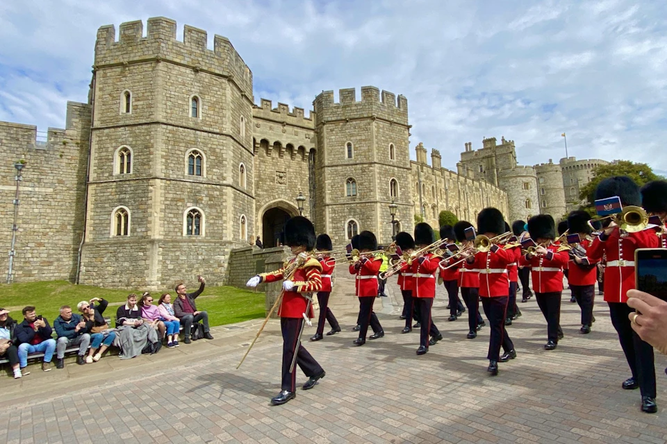A marching band in red uniforms and black hats parades outside an old stone building, while spectators watch from a bench.