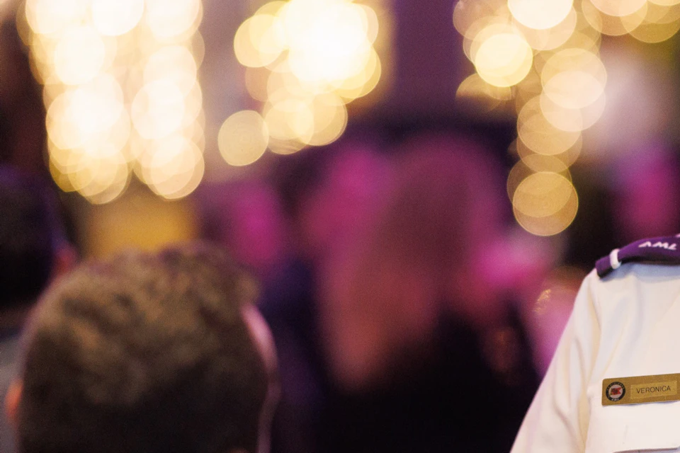 A server in uniform smiles while serving wine to a seated couple at a festive event with blurred lights in the background.