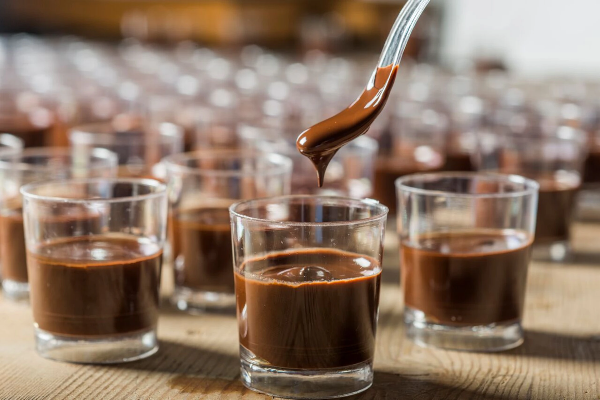 A spoon drips chocolate into a small glass, surrounded by numerous similar glasses filled with chocolate.