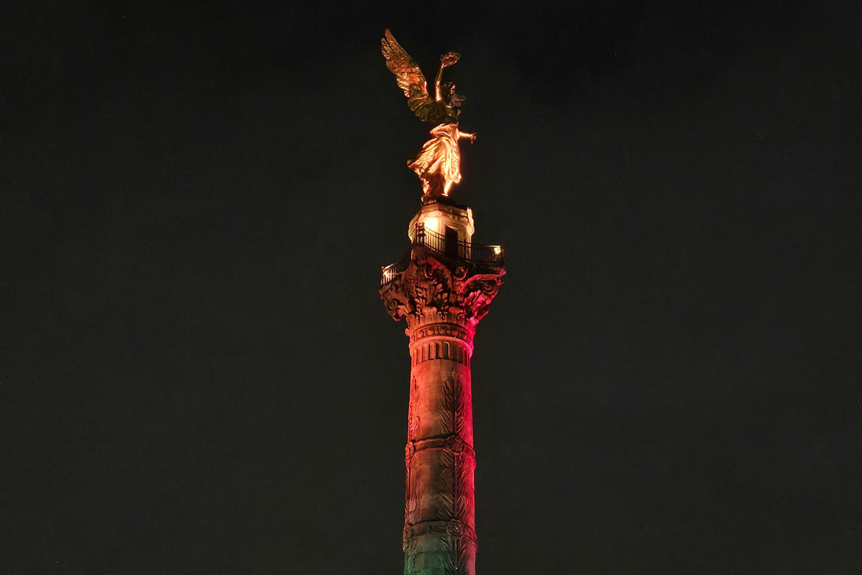 Een hoog zuilmonument met daarop een gouden engelfiguur, verlicht met rode en groene verlichting tegen een donkere nachtelijke hemel.