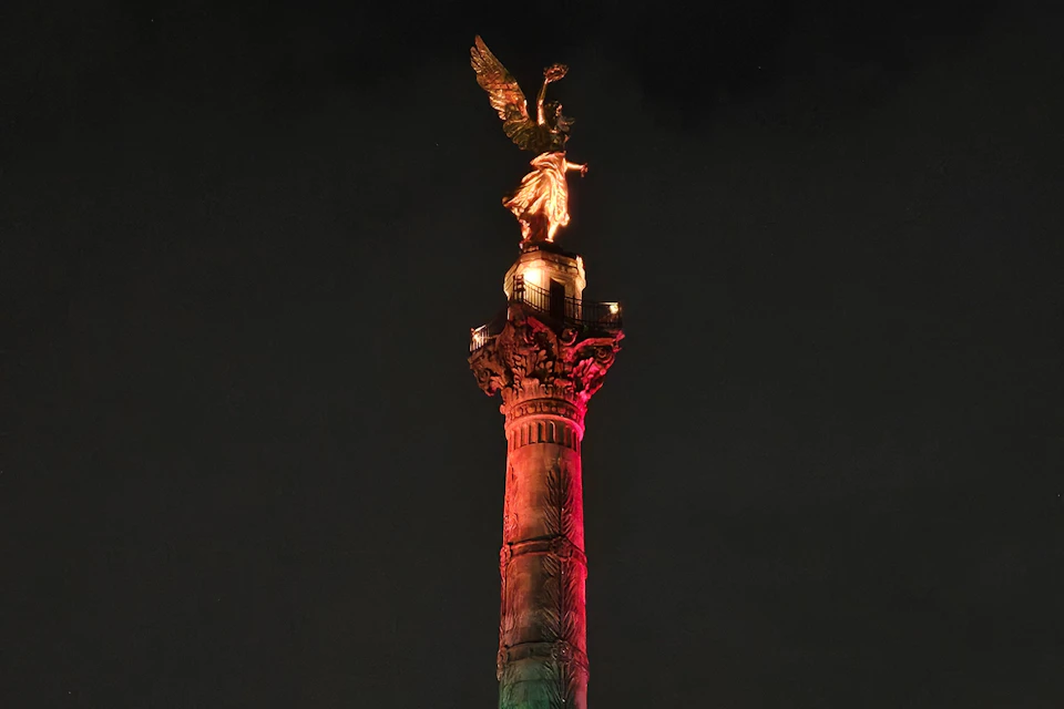 A tall column monument topped with a golden angel figure, illuminated with red and green lighting against a dark night sky.
