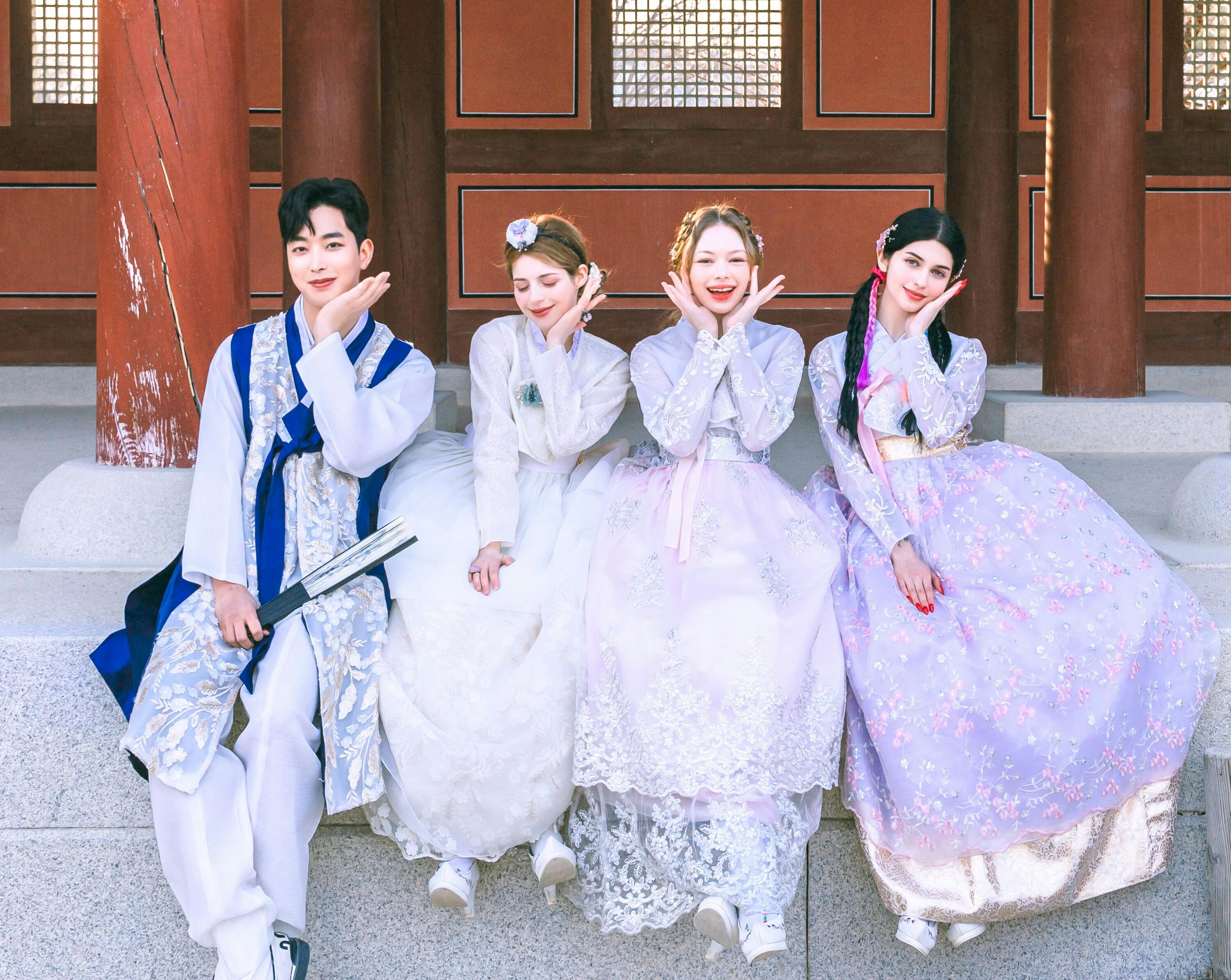 Four people in traditional Korean hanbok attire pose seated in front of a traditional wooden building, smiling cheerfully.
