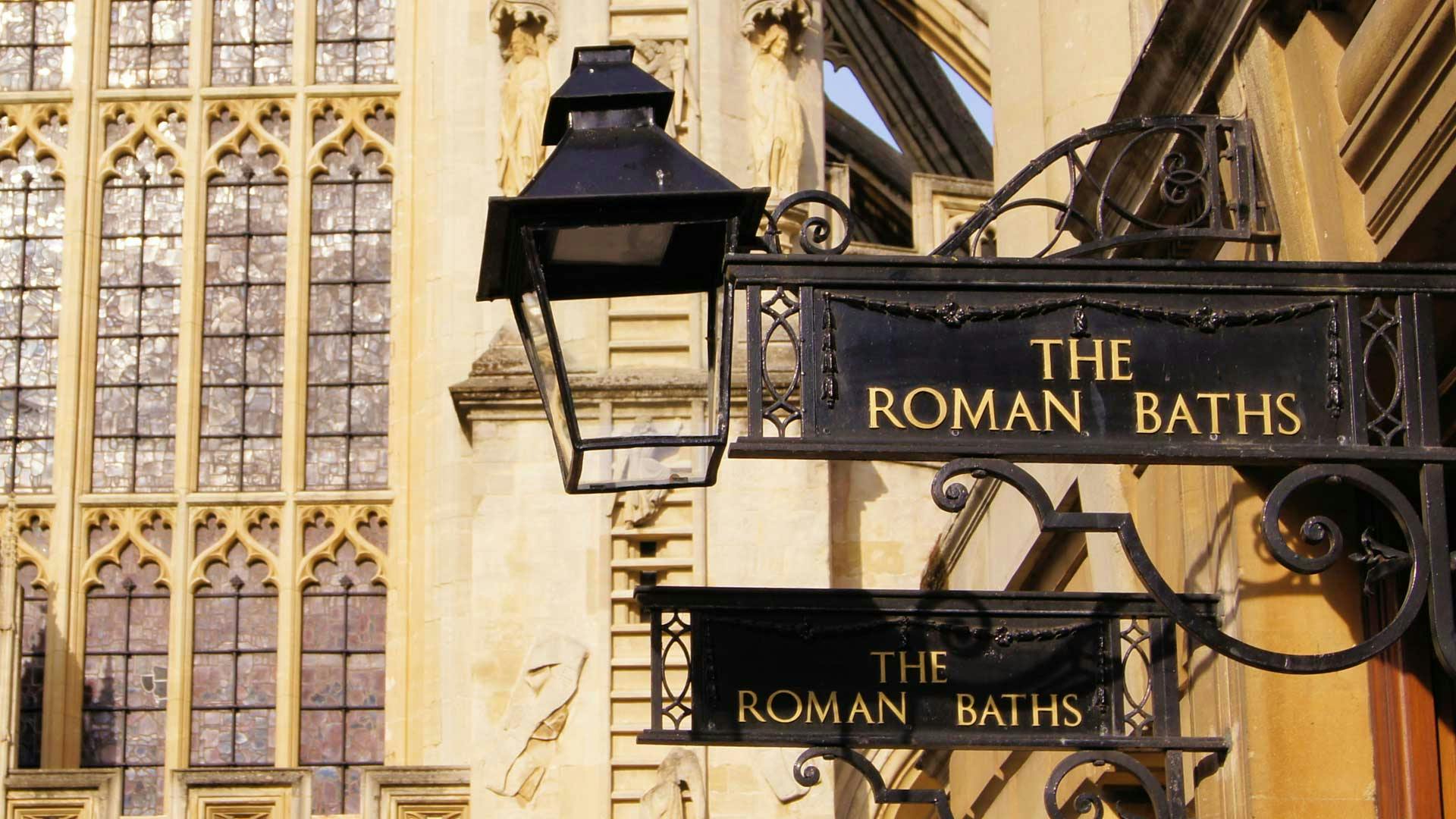Μαύρη επιγραφή με χρυσό κείμενο που γράφει «Τα Ρωμαϊκά Λουτρά» δίπλα σε ένα φανάρι, τοποθετημένο πάνω σε ένα ιστορικό πέτρινο κτίριο με αγάλματα.
