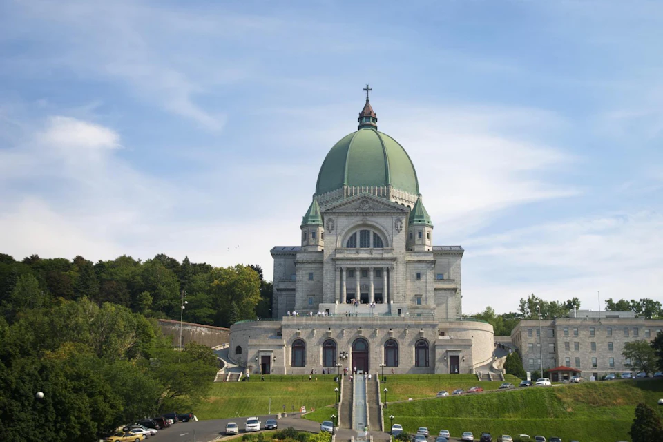 A large basilica with a green dome and cross, surrounded by trees, cars parked, and people walking up steps under a blue sky.