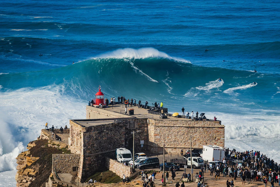 A large wave near a coastal fort with a red lighthouse, surrounded by spectators and vehicles.