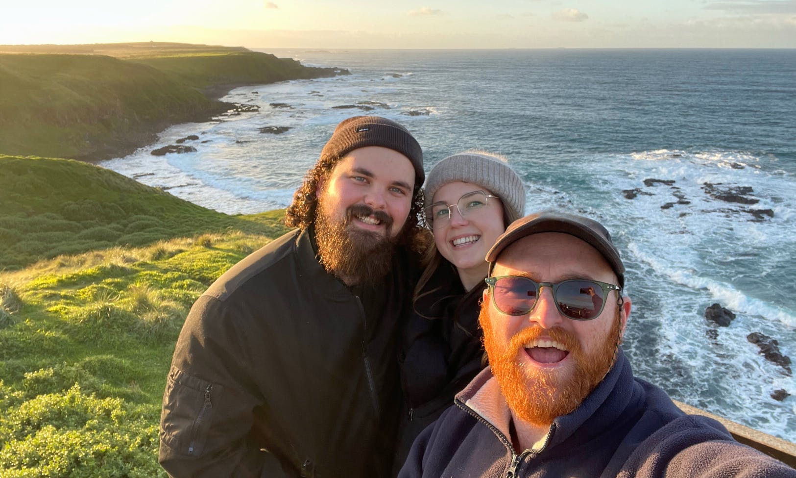 三个人在风景优美的海岸线上微笑，悬崖和海浪在一片多云的天空下。