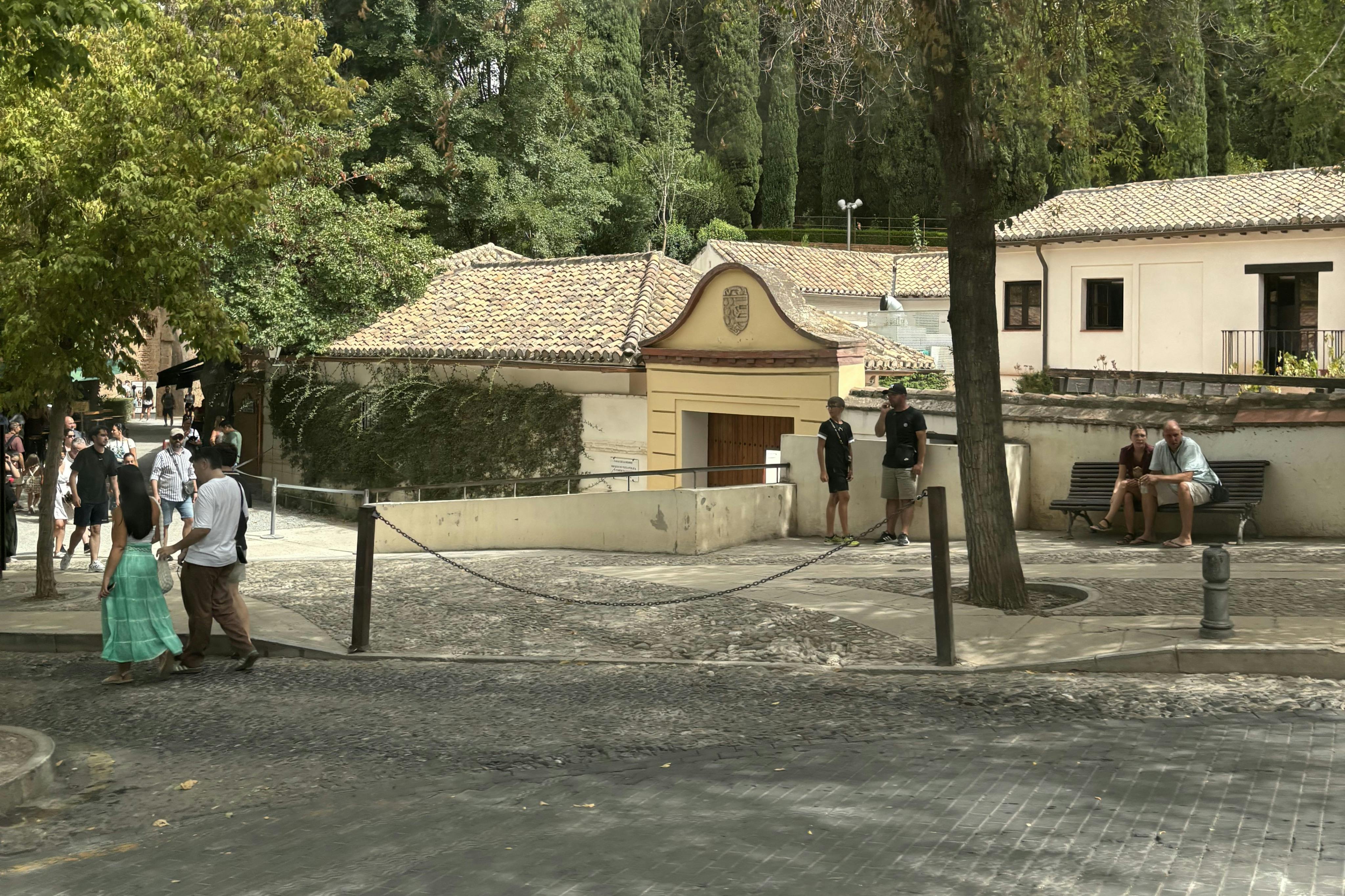 两人站在一栋建筑物的围栏入口附近，后面有瓷砖屋顶和树木。 旁边有一条人行道弯曲。