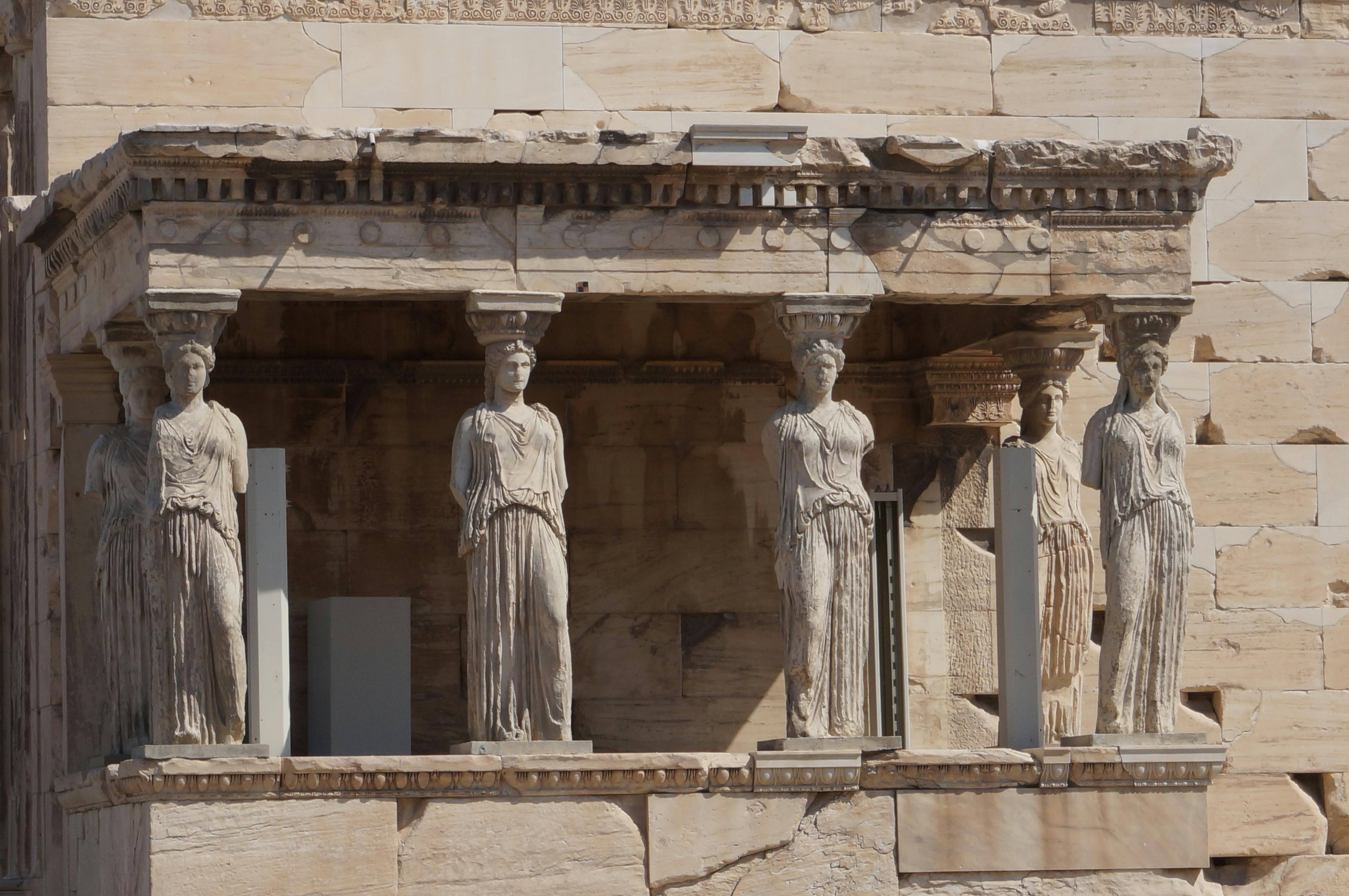 Three stone statues of robed women serve as columns on an ancient building facade with intricate carvings.