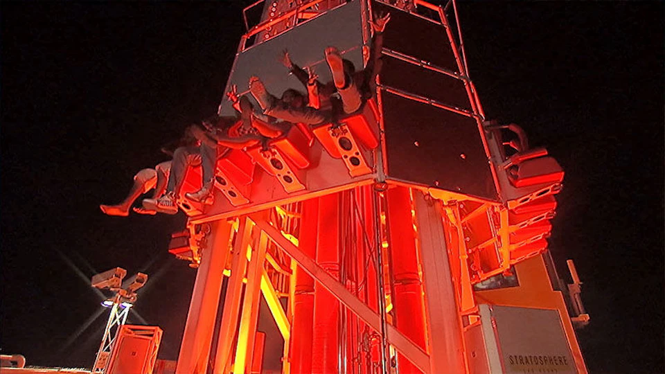 People on a drop tower ride at night, captured mid-descent, with bright red lighting illuminating the structure.