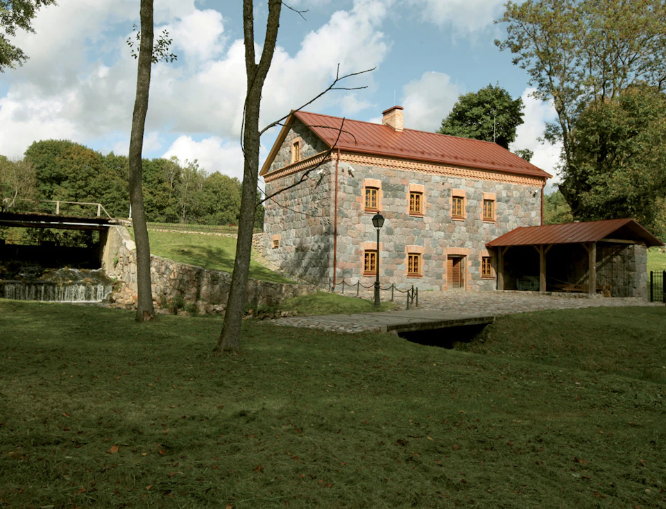 Stone house with a red roof surrounded by trees, a grassy lawn, a cobblestone path, and a small bridge over a stream.
