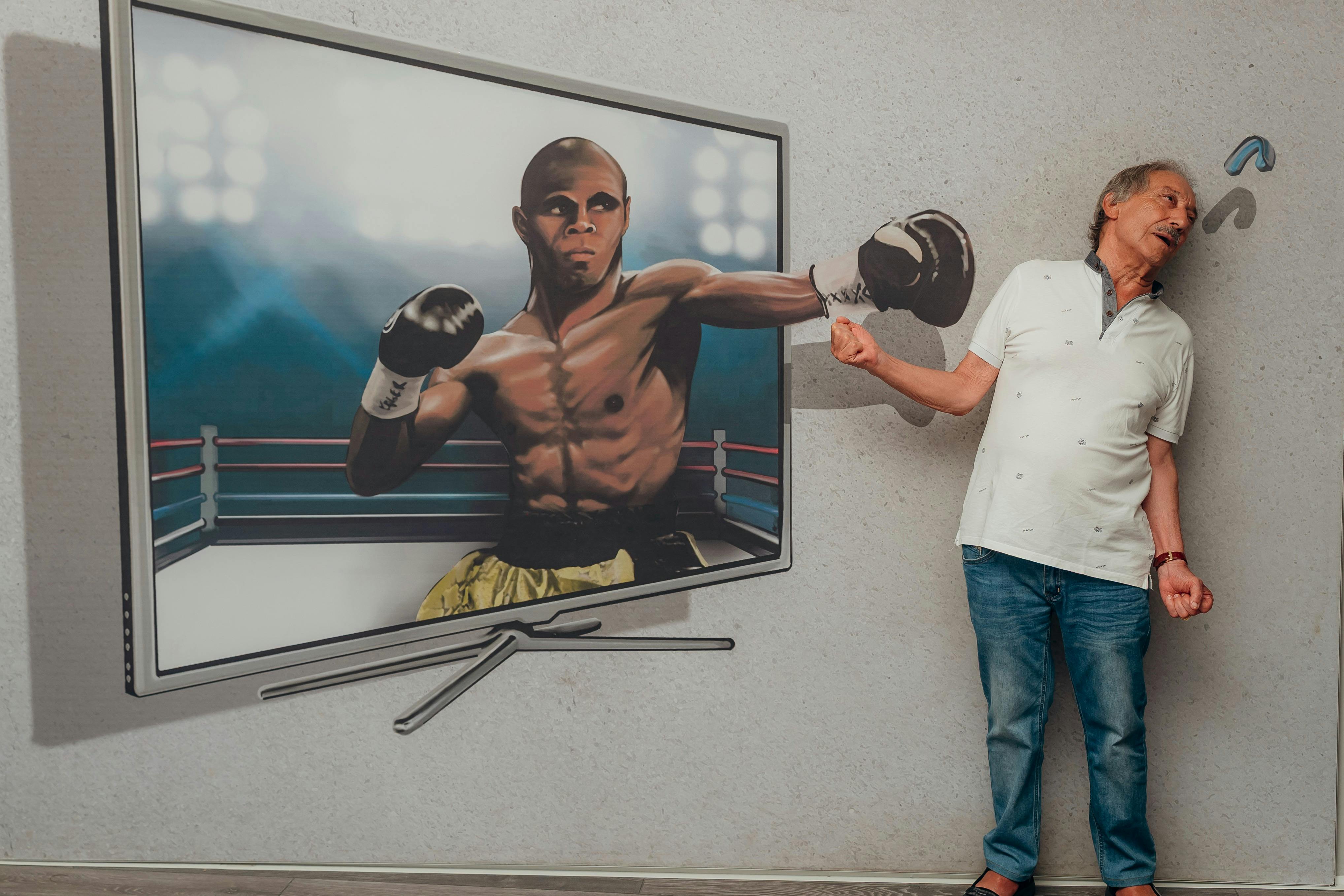Un homme en jeans et chemise blanche se tient à côté d'une peinture représentant un boxeur, semblant donner un coup de poing dans le gant étendu du boxeur.