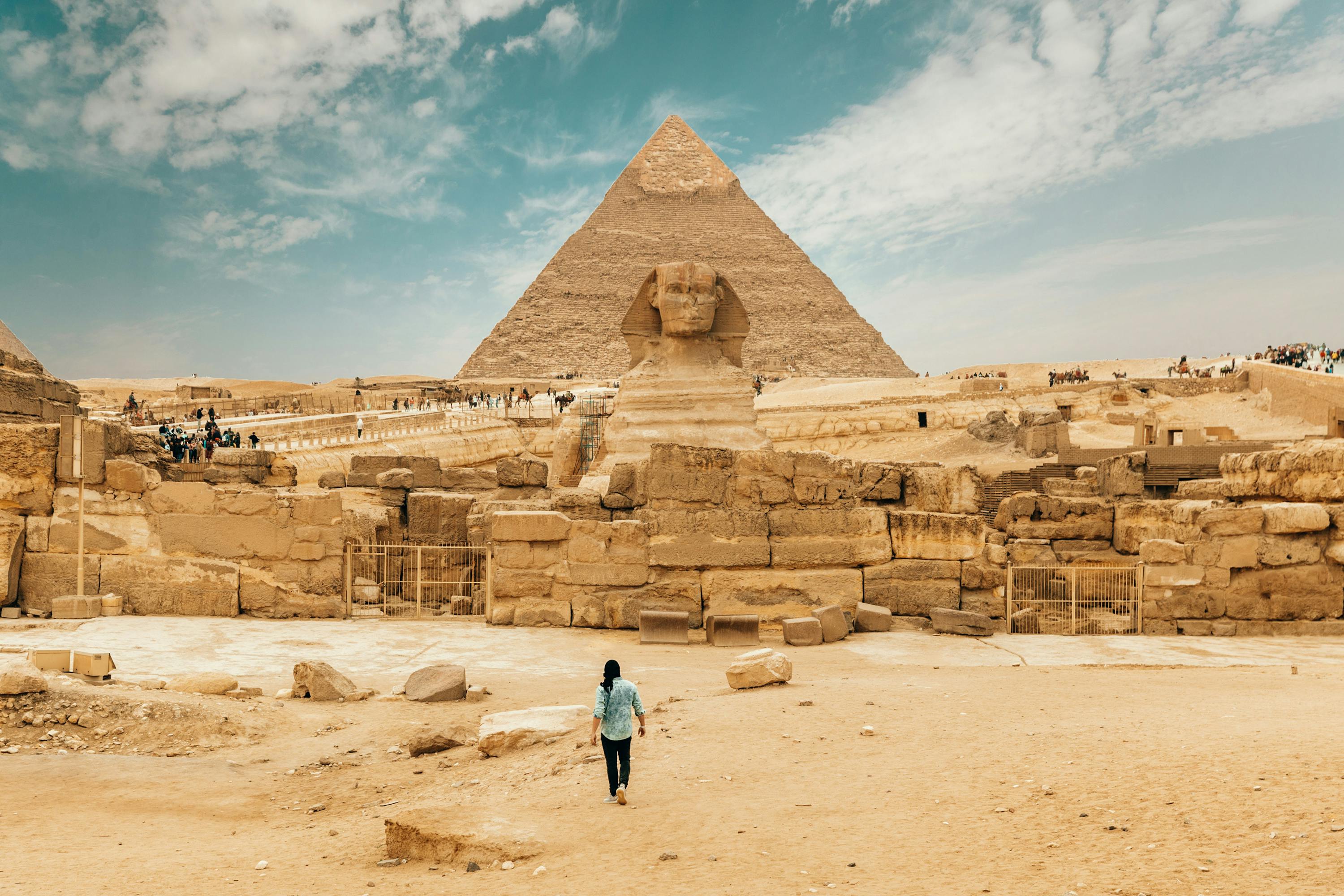 Une personne marche vers le Sphinx et une pyramide à Gizeh, avec un paysage désertique et des touristes éparpillés en arrière-plan.