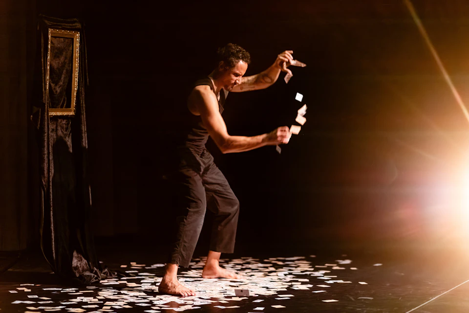 A performer on a dimly lit stage in a dramatic pose, surrounded by scattered cards on the floor.