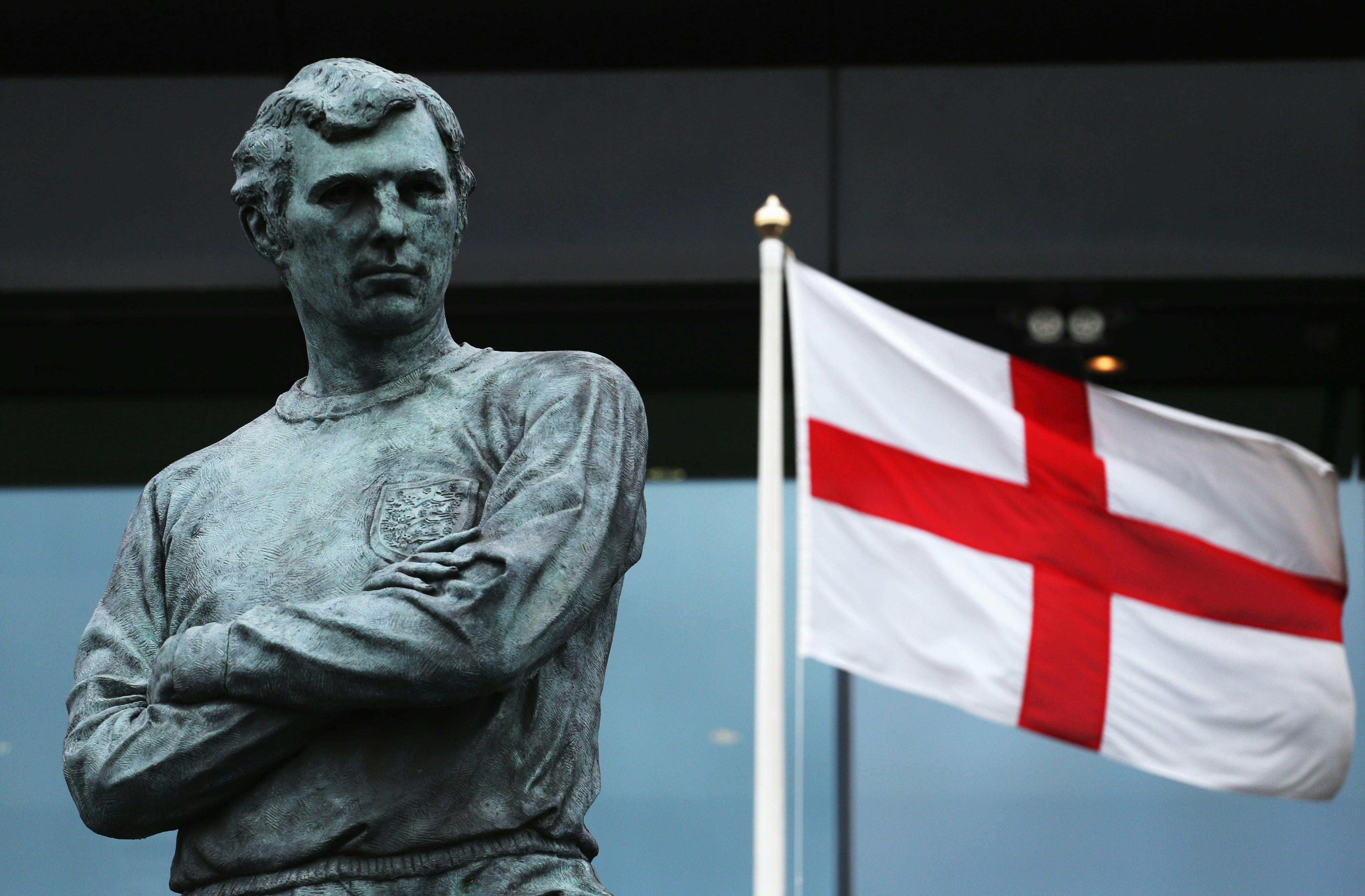 一尊穿着球衣、双臂交叉的男子铜像，旁边是一面旗帜，白色背景上有一个红十字。