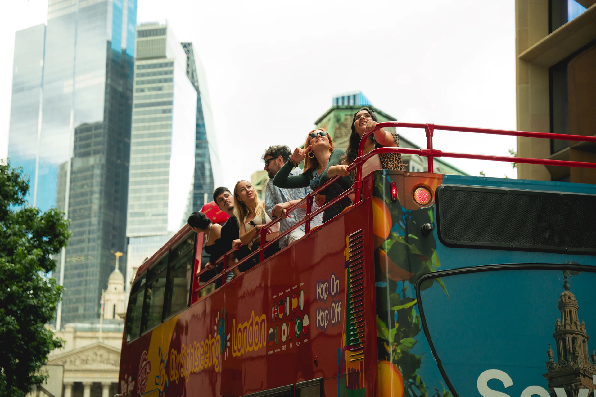 A group of people on the upper deck of a red sightseeing bus in a city with modern skyscrapers and older architecture.