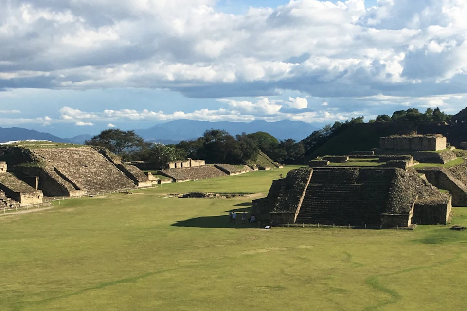 Ancient stone pyramids and platforms surrounded by green grass and trees under a partly cloudy sky.