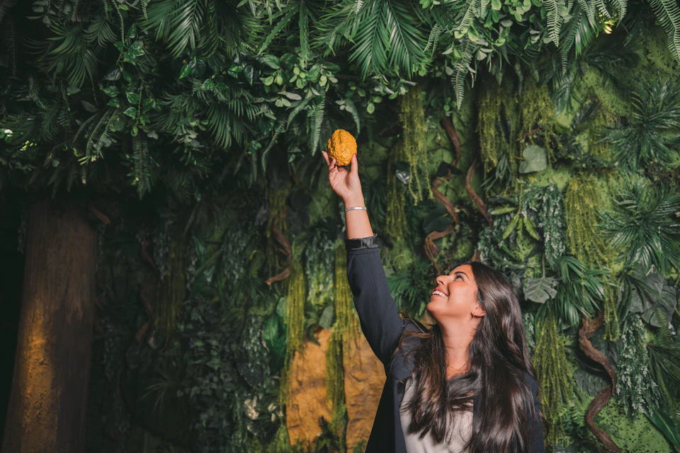 A woman reaches up smiling, holding a yellow object, against a lush green background of leaves and moss.