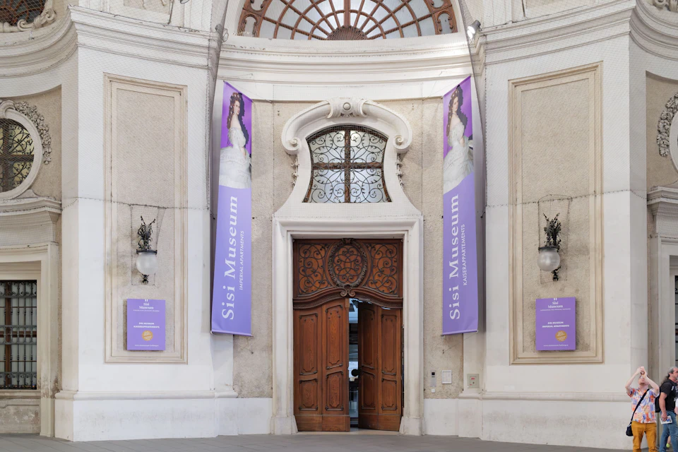 Entrance to the Sisi Museum, featuring large wooden doors, arched windows, and purple banners displaying an image of Empress Sisi.